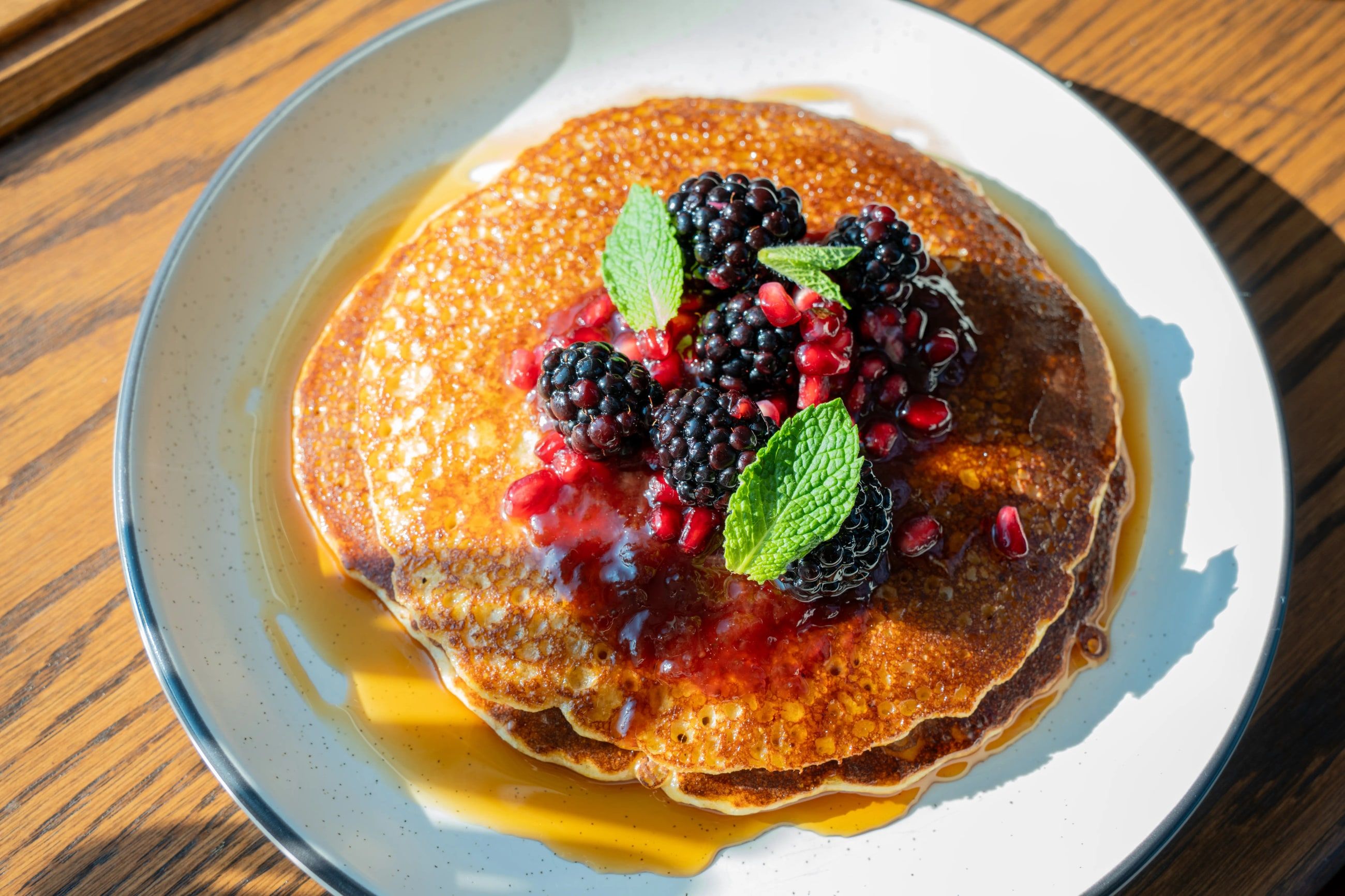A stack of pancakes with fresh berries, mint and maple syrup on a wooden table in the sun.